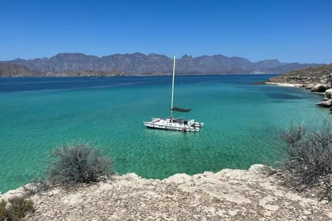 Sailboat on clear turquoise water near rocky shore, distant mountains in background.