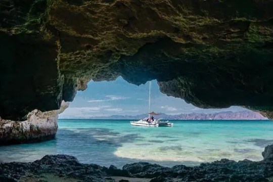 View from cave opening of a sailboat on turquoise water with distant mountains.