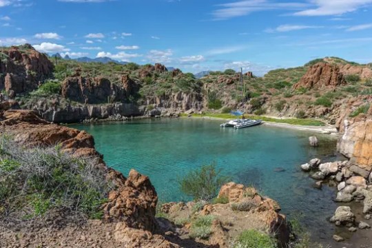 Scenic cove with clear turquoise water, rocky cliffs, and a small dock under a blue sky with clouds.