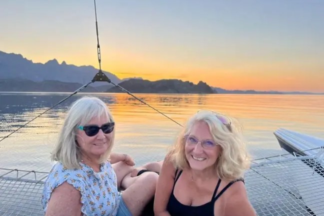 Two women smiling on a boat net at sunset with mountains in the background.