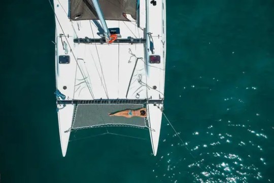 Aerial view of a person relaxing on a catamaran's net over turquoise water.