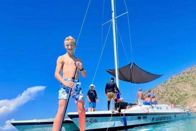 Boy paddleboarding near a sailboat on clear blue water under a sunny sky.