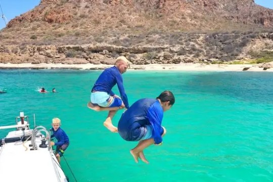 Two kids in swimwear jump off a boat into turquoise water.