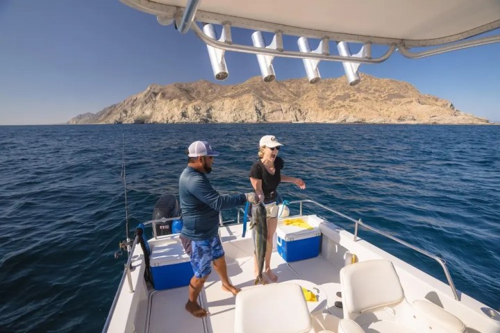 Two people on a boat holding a large fish near a rocky coastline.