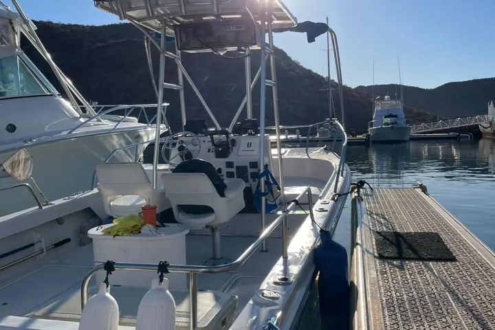 Fishing boat docked at marina under bright sun with fishing rods and equipment visible.