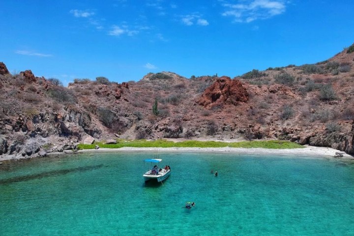 Boat in clear turquoise water near rocky island with shrubs under a blue sky.