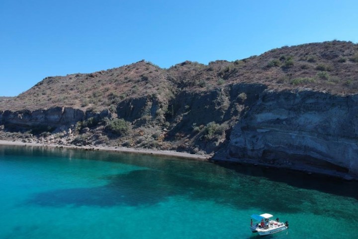 Small boat on turquoise water near rocky hillside under clear blue sky.